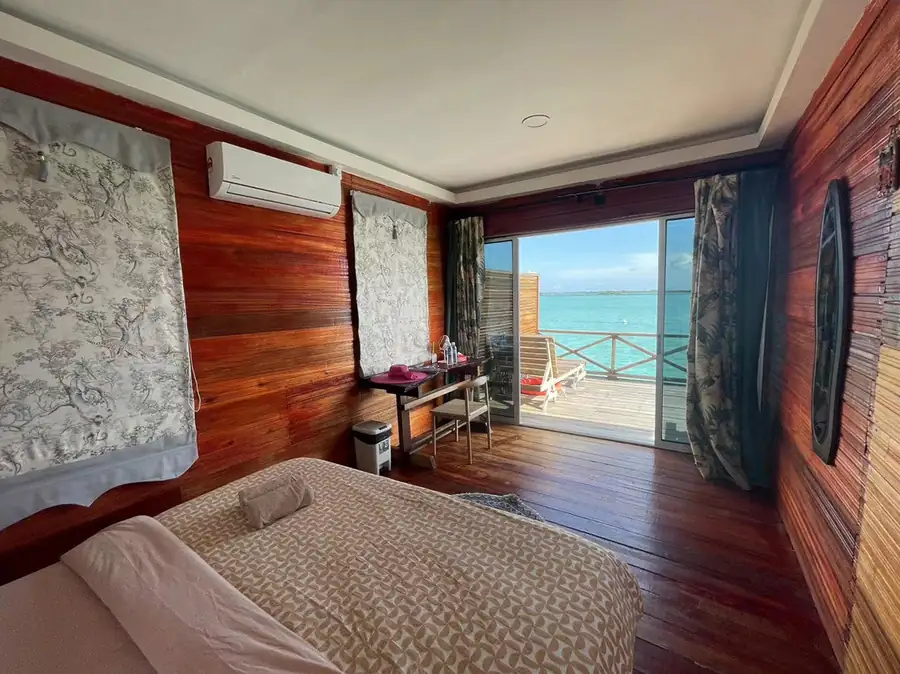 A warm bedroom at Fairyland Resort near the Tun Sakaran Marine Park focusing on a cozy bed with patterned linens, a vibrant pink floral painting, rustic timber floor with smooth boards, and A/C.
