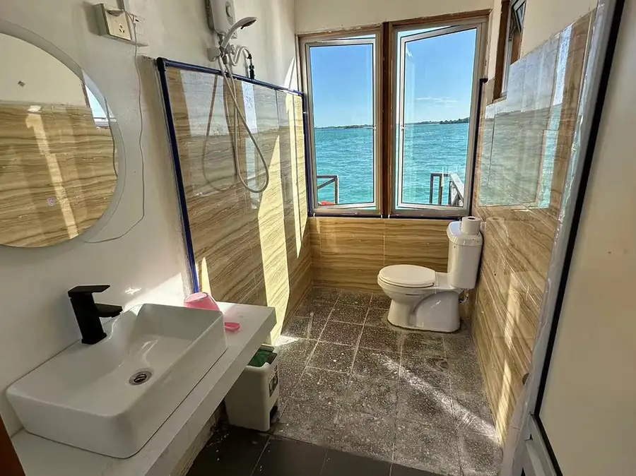 A cozy bathroom at Fairyland Resort detailing warm wood-paneled walls, a modern washbasin cabinet, a round mirror, a classic white toilet, and an open shower area with an electric shower.