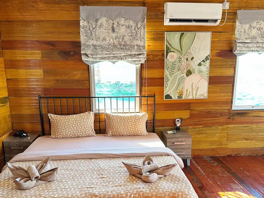 A tranquil bedroom at Fairyland Resort near Bum Bum Island focusing on a cozy bed with white linens, a wooden bedside table with a lamp, smooth varnished timber flooring, and AC.