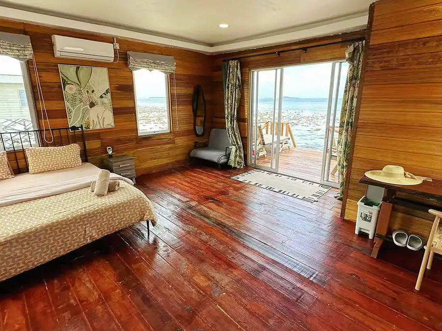 An inviting bedroom at Fairyland Resort in Semporna, featuring a bed facing panoramic glass doors to a private deck, rustic timber walls, smooth wooden boards, modern furniture, and air conditioning.