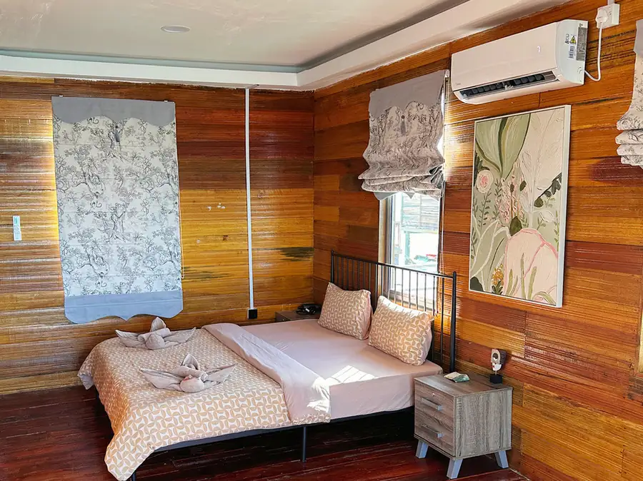 A romantic bedroom at Fairyland Resort over the water, presenting a plush bed with swan towels, botanical artwork, elegant wooden plank walls, rich varnished timber flooring, and quiet air con.