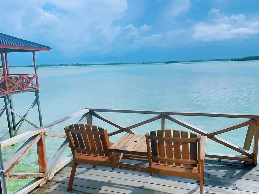 Private sun-soaked balcony featuring a wooden bench seat at Fairyland Resort. An overwater sanctuary suspended over the Celebes Sea, perfect for admiring the crystal-clear waters.