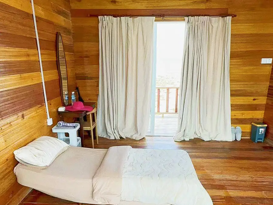 A cozy single bedroom at Fairyland Resort over the water, offering a bed facing panoramic glass doors to the balcony, warm wooden plank walls, varnished timber flooring, a vanity desk, and AC.