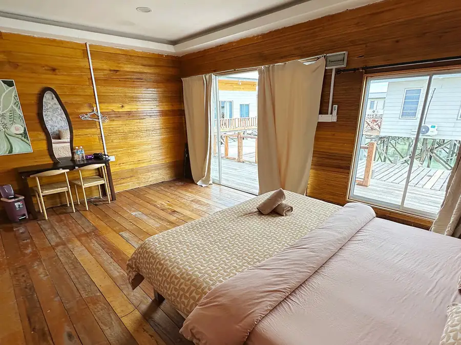 A bright bedroom at Fairyland Resort near Pulau Bum Bum featuring a bed facing glass doors opening onto the balcony with seaviews, rustic wood walls with glossy floors, and AC.