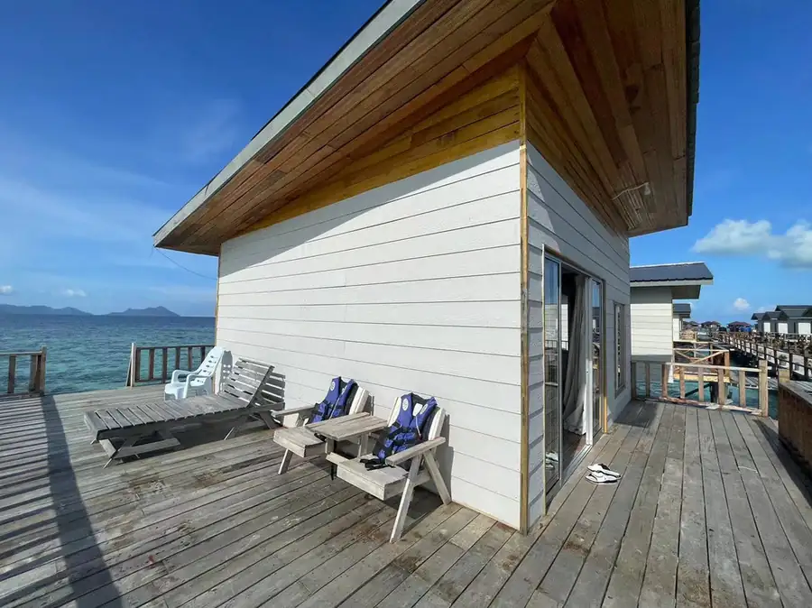 A traditional Sabah style timber chalet featuring a prominent pointed roof, glass sliding doors, and a spacious overwater wooden deck with a hammock net at Fairyland Resort.