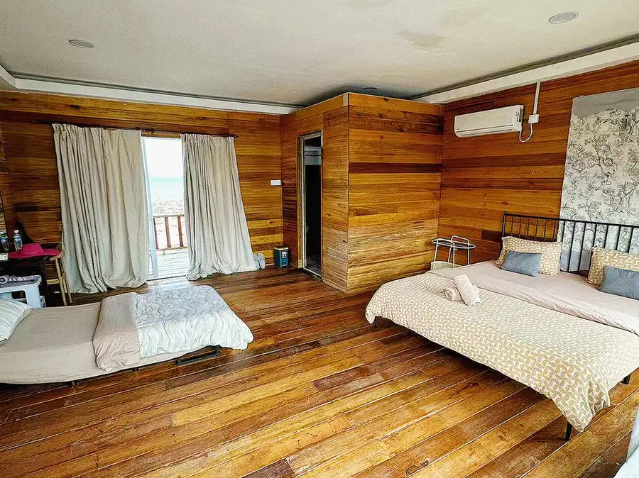 A large bedroom at Fairyland Resort in Semporna featuring two beds facing panoramic glass-panel doors framing the seaviews, traditional Sabahan style wood paneling with rustic floors, and air con.