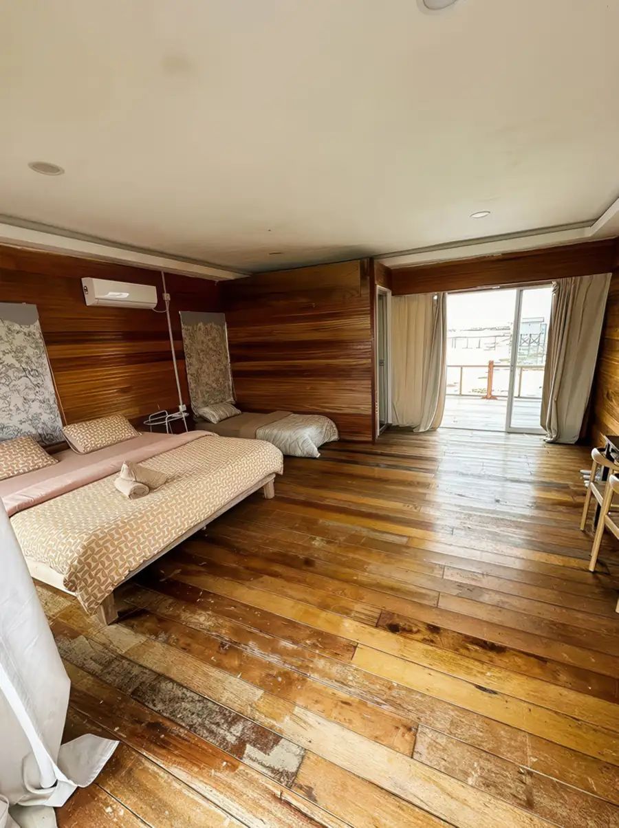 A generous triple bedroom at Fairyland Resort looking at Bohey Dulang, featuring beds facing glass-panel doors to a balcony, traditional Sabahan style wood paneling, gleaming timber floors, and A/C.